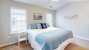 Quarto com cama, duas janelas, ventilador de teto e obras de arte nas paredes.