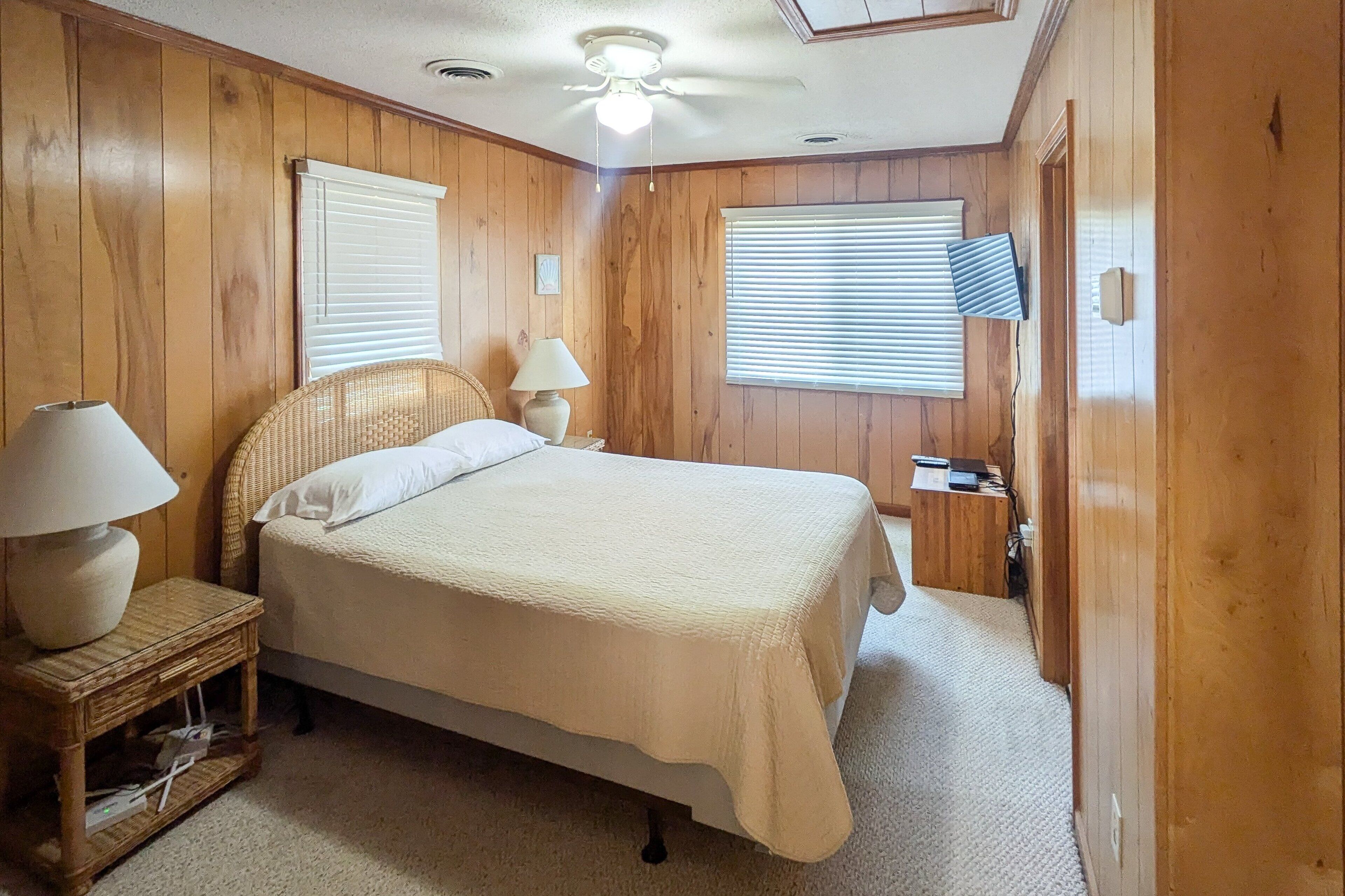 Une chambre avec des lambris en bois, un lit recouvert d’une courtepointe blanche, une table de chevet avec une lampe et un ventilateur de plafond.
