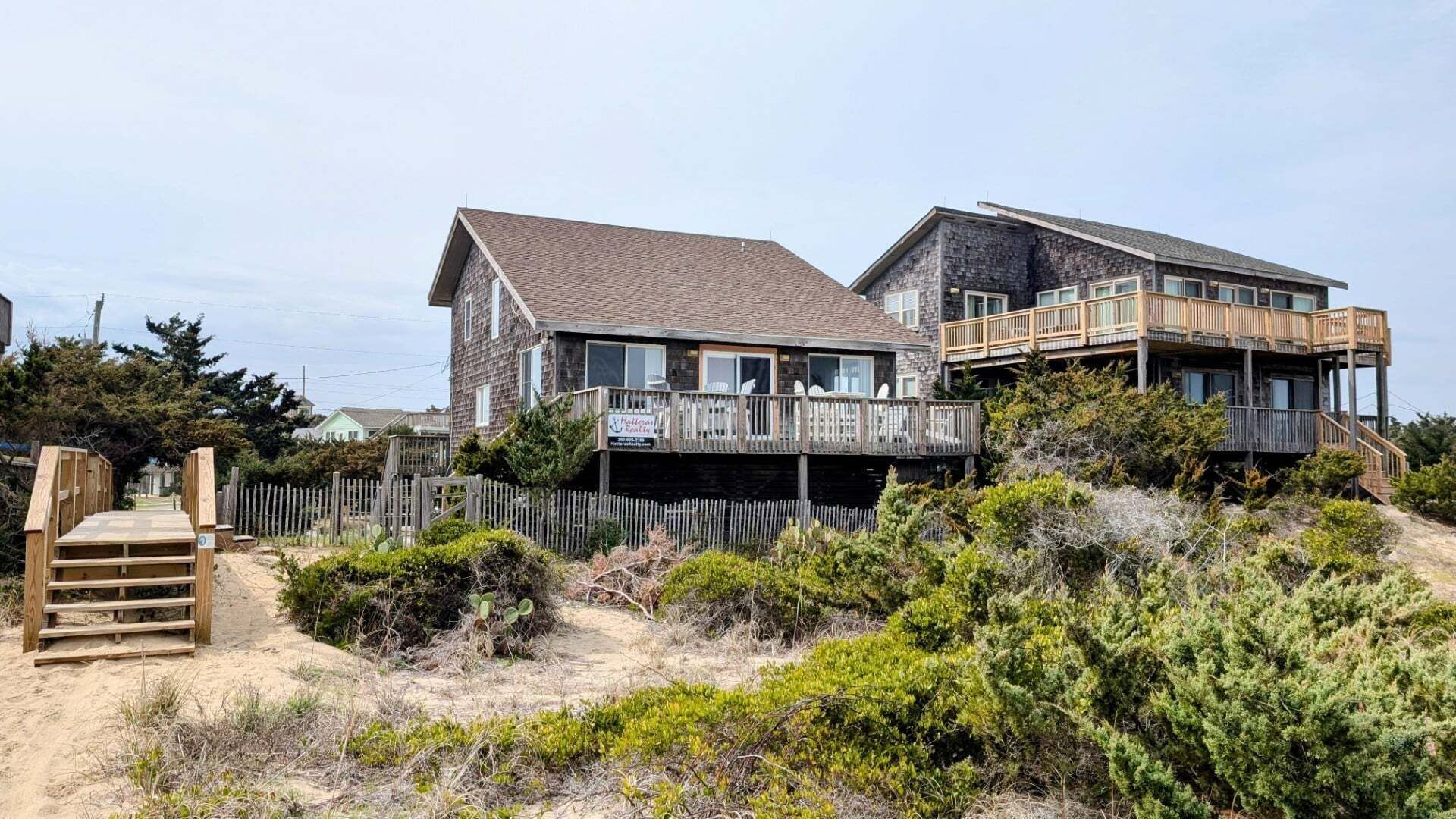 A two-story beachfront property with a wooden deck, a staircase leading to the entrance, and a sandy foreground with vegetation.