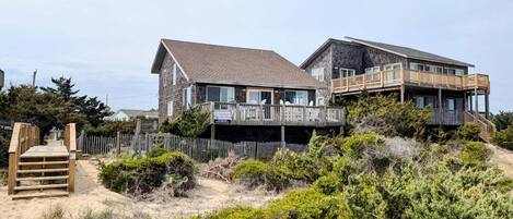 A two-story beachfront property with a wooden deck, a staircase leading to the entrance, and a sandy foreground with vegetation.