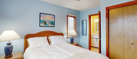 Un dormitorio con cama, dos mesitas de noche con lámparas, un ventilador de techo, una ventana con persianas, una puerta y un cuadro en la pared.