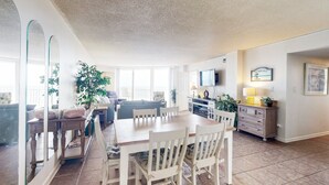 Amplia sala de estar con una mesa de comedor, sillas, sofá y televisor. Hay plantas y un mueble con cajones.