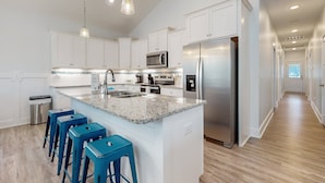 A modern kitchen with white cabinetry, a stainless steel refrigerator, a microwave, and a granite countertop. There are three blue bar stools and a trash can. - Knee Deep: Family friendly, near beach (Oak Island)
