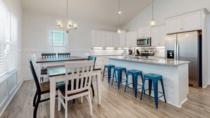A modern kitchen with white cabinetry, a central island with bar stools, a stainless steel refrigerator, and a dining area with a wooden table and chairs. - Knee Deep: Family friendly, near beach (Oak Island)