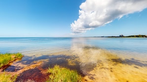 A calm beach with clear water, a grassy foreground, and a distant house on the horizon. - Waterfront House w/ Sound View, Shared Pool, Private W/D, Private Hot Tub (Frisco)