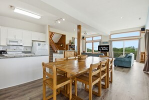 A modern kitchen with white cabinets, a dining area with a wooden table and chairs, a staircase, and a living room with a blue sofa and large windows. - Waterfront House w/ Sound View, Shared Pool, Private W/D, Private Hot Tub (Frisco)
