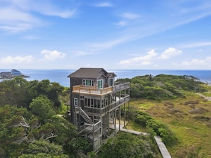Une maison située sur la côte, dotée d’une terrasse en bois et de plusieurs niveaux, entourée d’une végétation luxuriante et offrant une vue sur la mer.