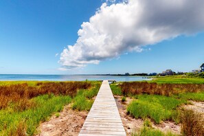 A wooden boardwalk leading to a beach with grassy banks and houses in the background.