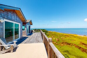 A wooden deck with a view of the ocean, a glass door entrance, and a bench. - Waterfront House w/ Sound View, Shared Pool, Private W/D, Private Hot Tub (Frisco)