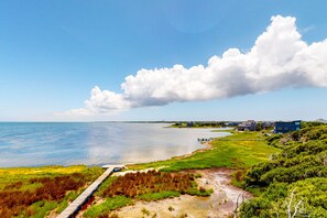 A coastal landscape with a wooden boardwalk leading to a beach, houses in the background, and a large cloud in the sky.