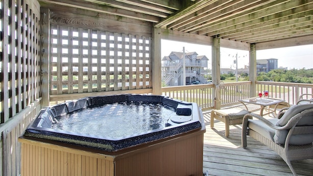 Une terrasse en bois avec un jacuzzi, des meubles en osier et une vue sur une maison au loin.
