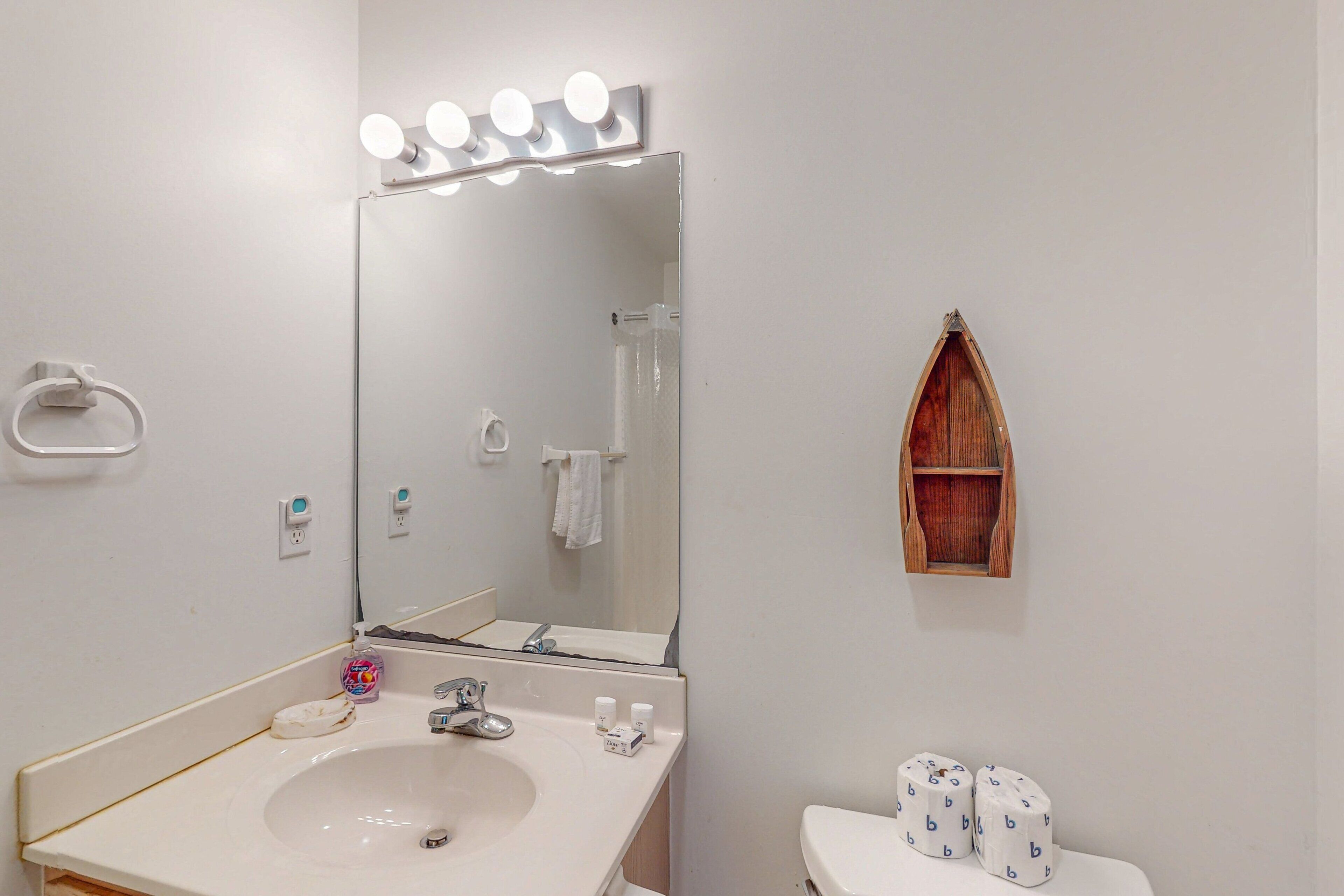 Een badkamer met een wastafel, spiegel, verlichting boven de spiegel, een handdoekhouder en een houten plank.