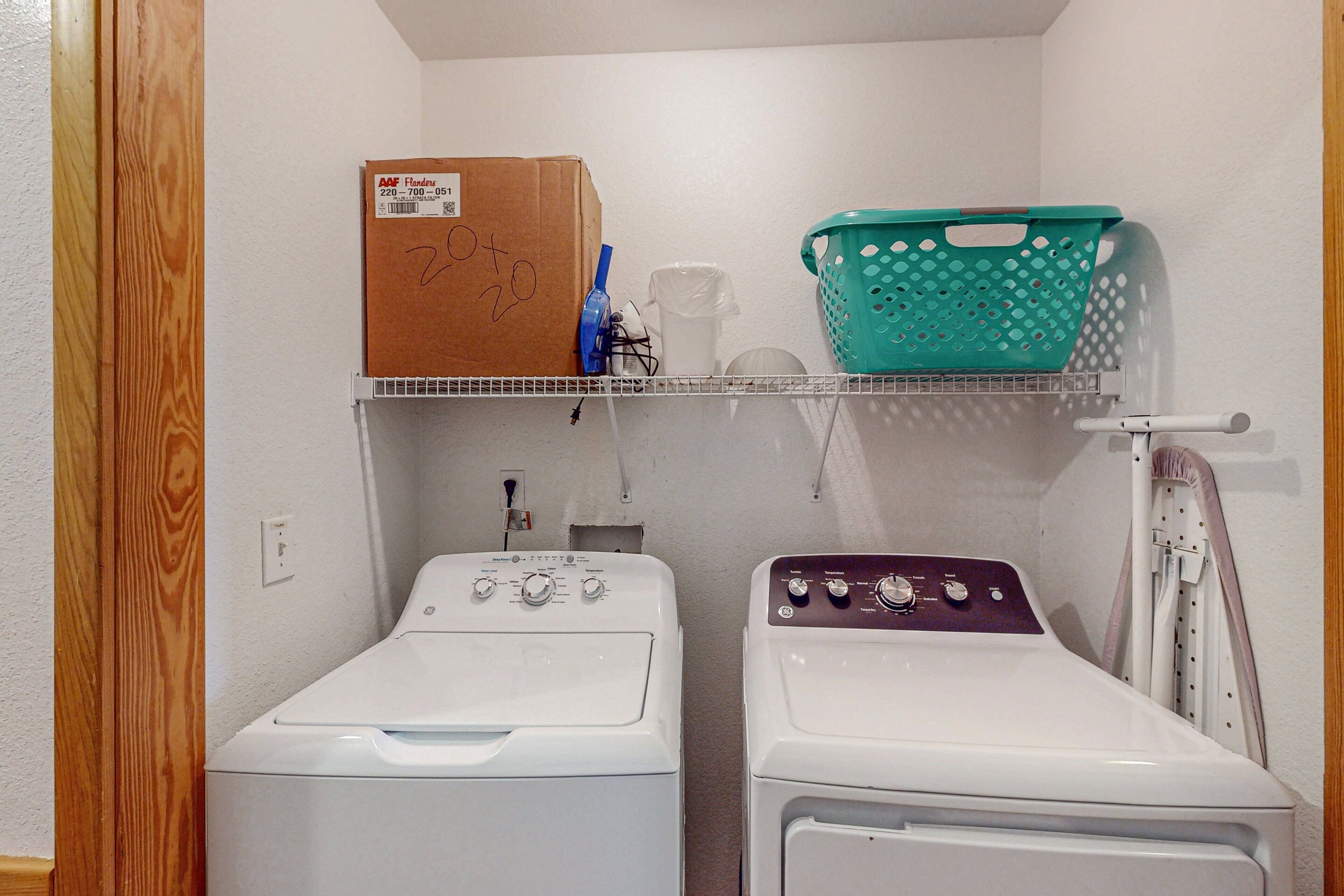 A laundry room with two washing machines, a shelf with a box and a basket, and a wall-mounted drying rack.