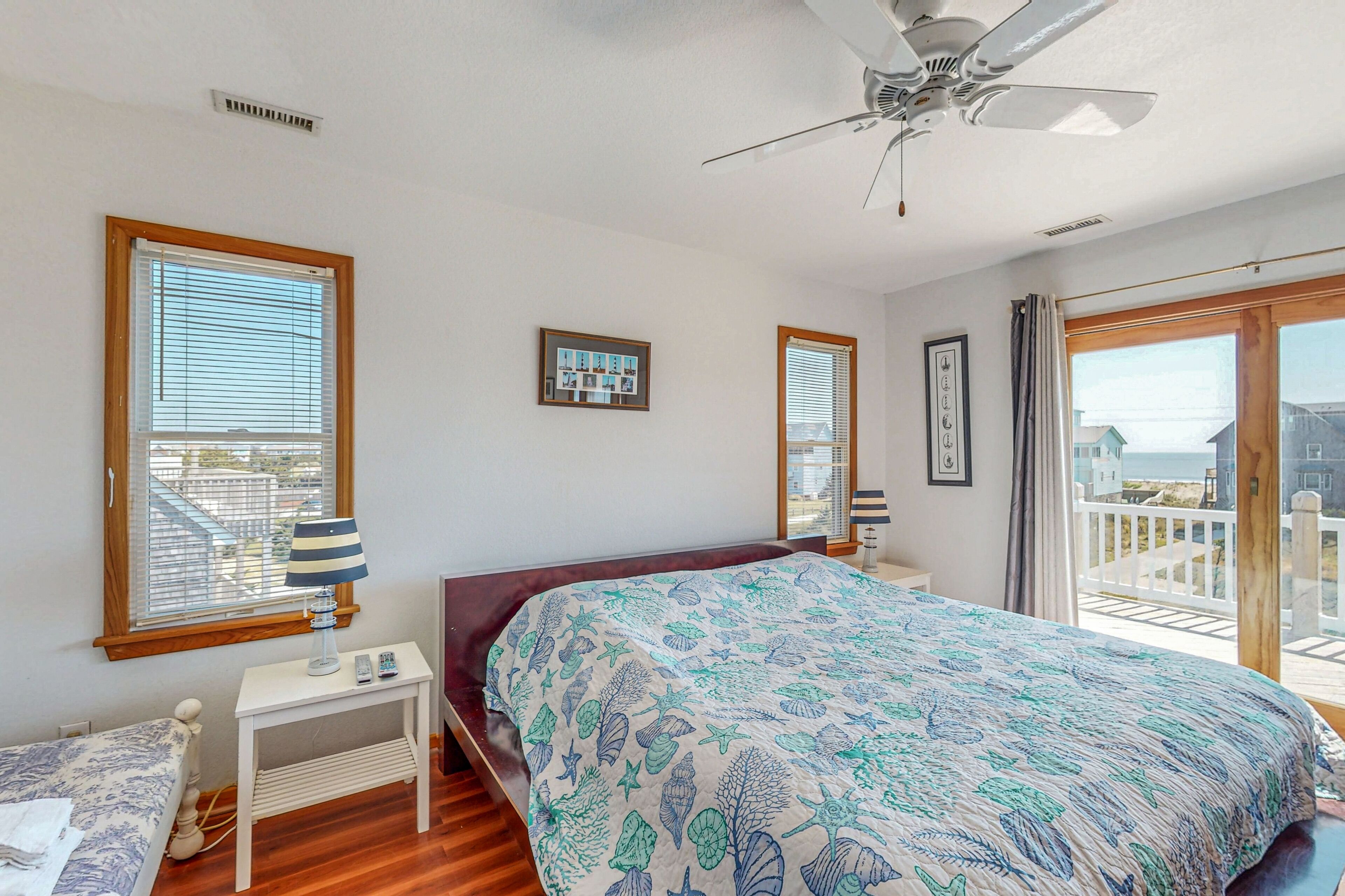 A bedroom with a bed, a bedside table with a lamp, a ceiling fan, and a view of the outside through a large window.