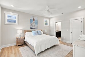 A bedroom with a bed, nightstands, a ceiling fan, a painting, and a bathroom visible through an open door.
