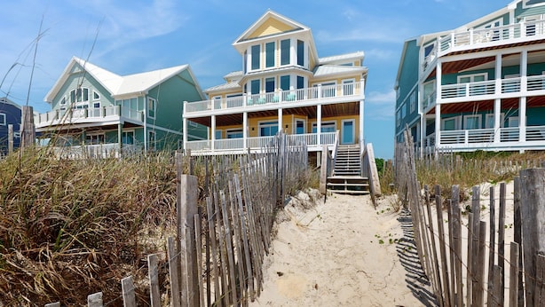 A beachfront property with multiple levels, a prominent balcony, and a sandy pathway leading to the entrance. - Oceanfront, multi-level home with beach access, views, game room, & elevator (North Topsail Beach)