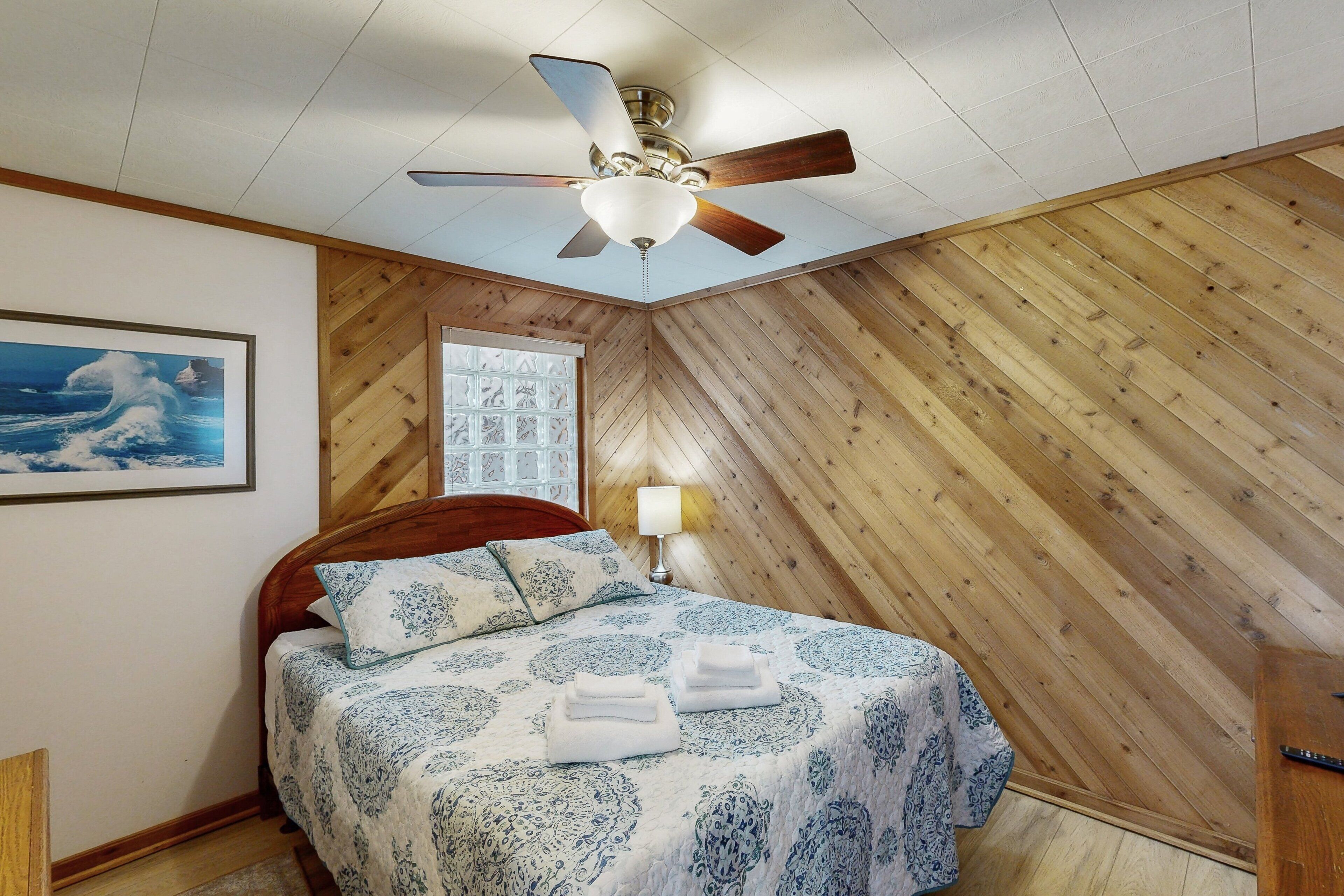 Une chambre avec un plafond en bois, un lit recouvert d’une courtepointe à motifs, un ventilateur de plafond et une fenêtre avec des stores.
