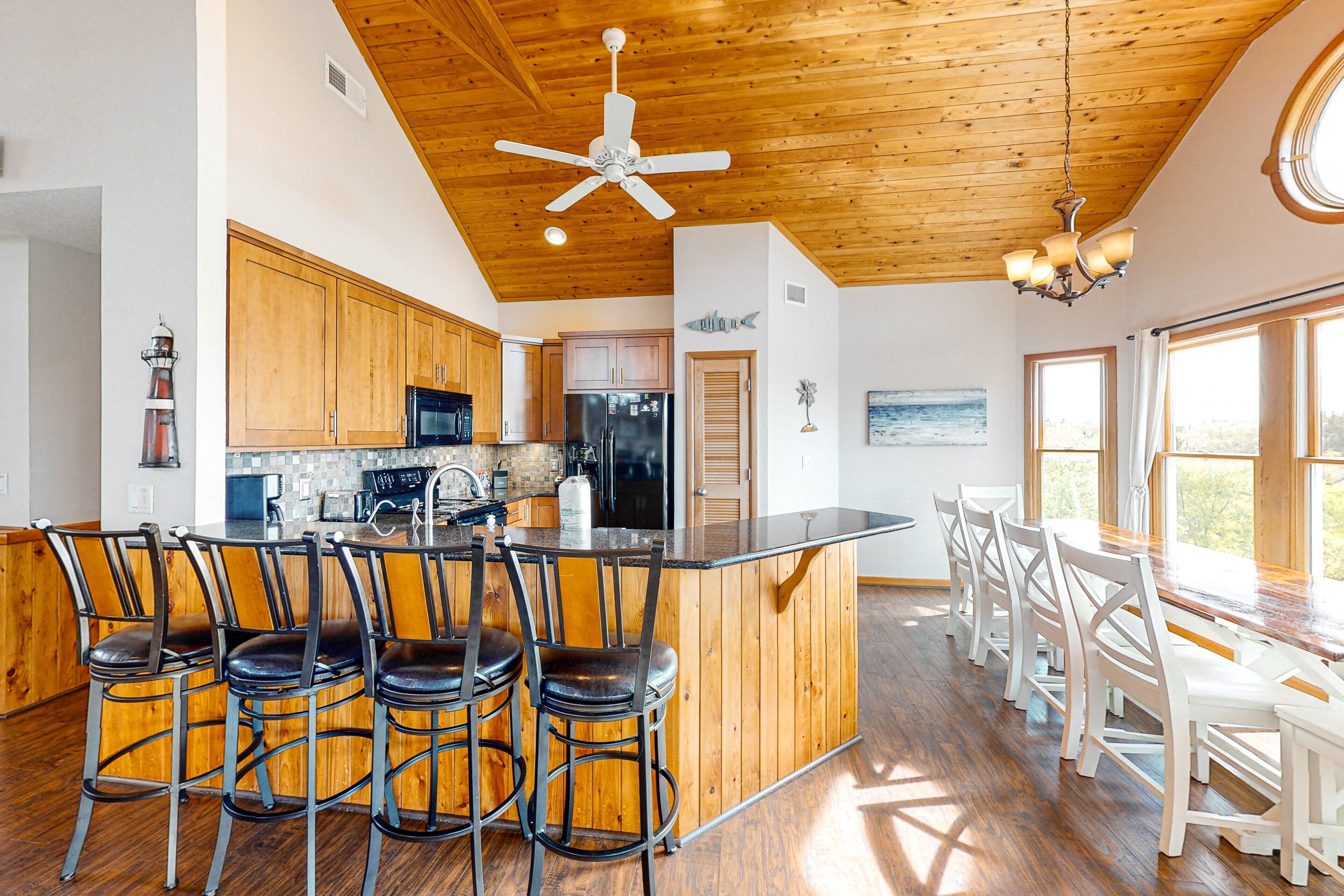 A kitchen with wooden cabinets, a central island with bar stools, a ceiling fan, and a dining area with white chairs.