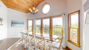 A dining area with a wooden table and white chairs, large windows with curtains, and a chandelier. - Reel 'Em Inn 403: Gorgeous, outdoor pool, spacious carport, decks (Avon)