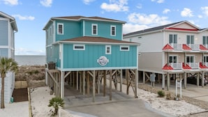 A beachfront property with a two-story house elevated on stilts, a separate building with a balcony, and a sandy beach area. - Wave Watcher: Stunning ocean front home with private hot tub and direct beach access (Oak Island)