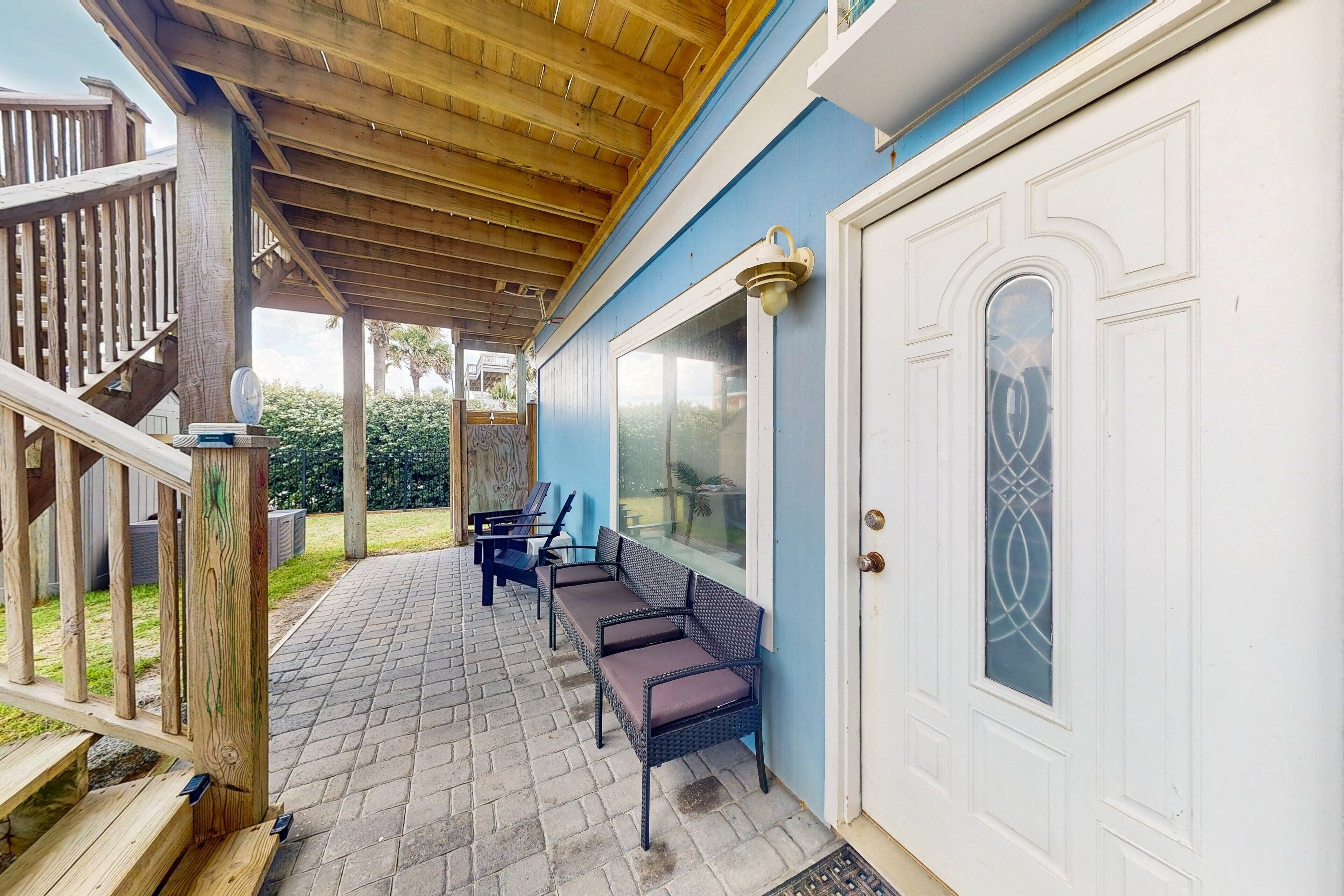 A covered patio with wooden flooring, a white door with a glass panel, and two wicker chairs.