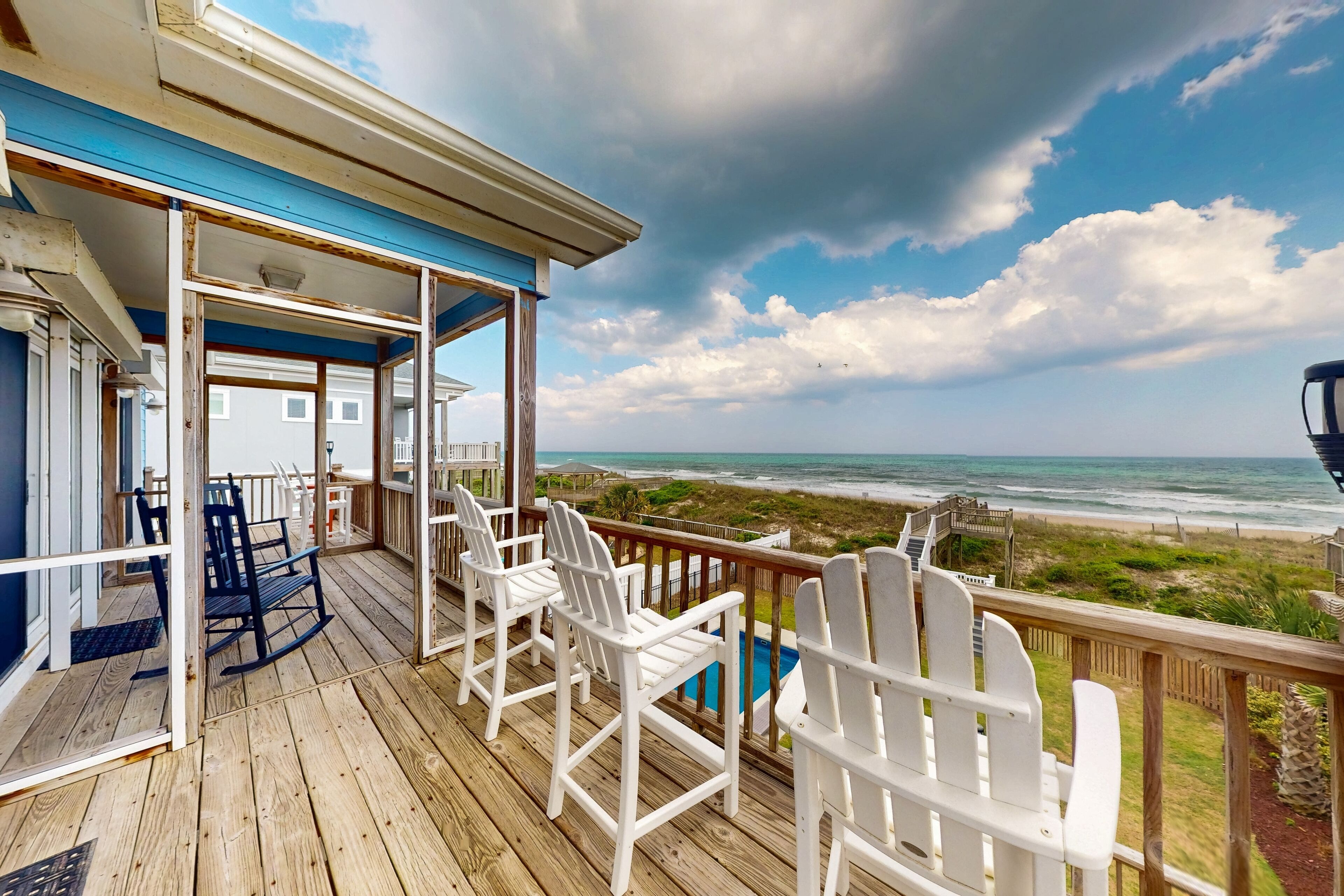 A wooden deck with white Adirondack chairs, a pool, and a view of the beach and ocean.