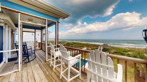 A wooden deck with white Adirondack chairs, a pool, and a view of the beach and ocean.