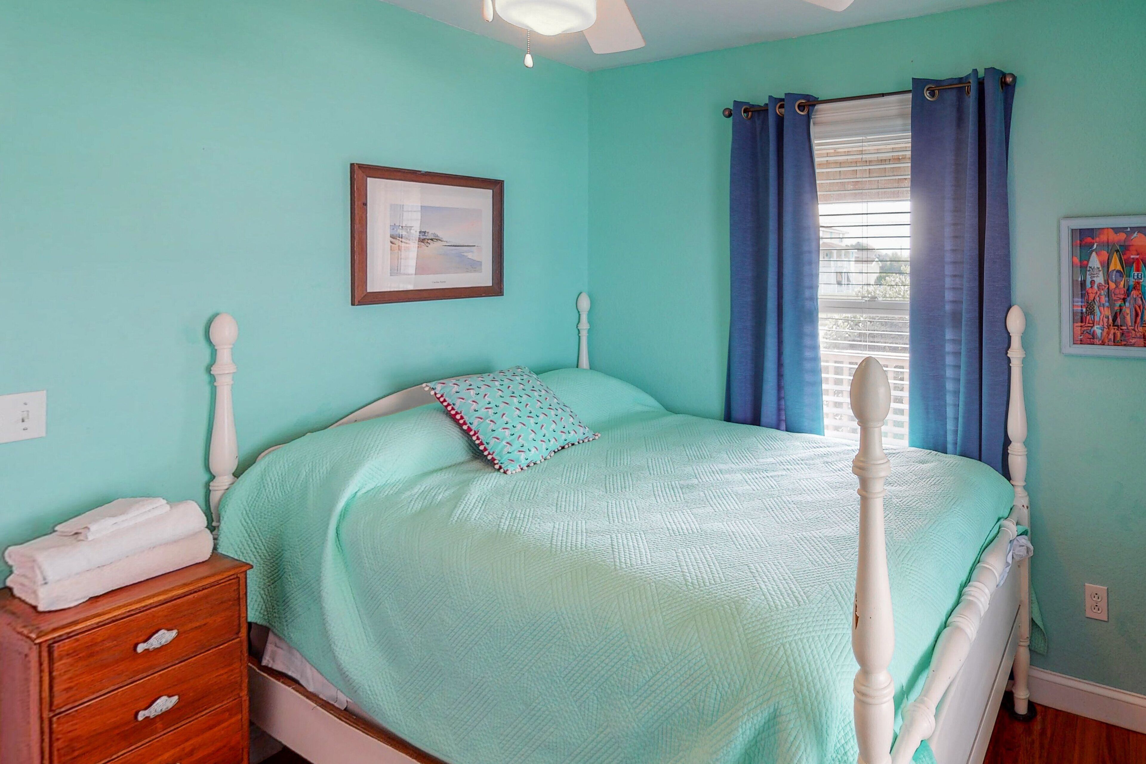 A bedroom with a bed, a nightstand, a framed picture, and a window with curtains.