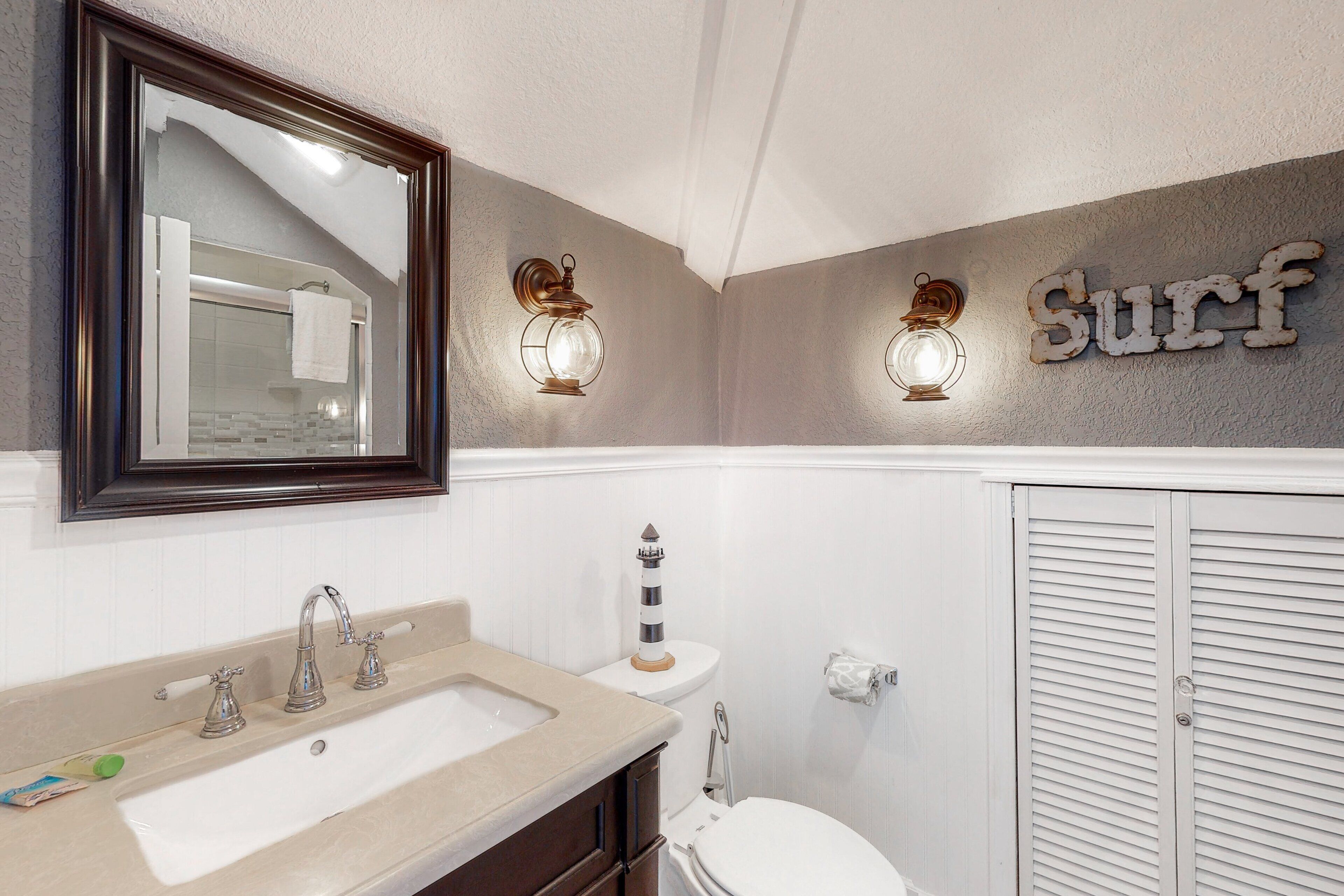 Une salle de bain avec un lavabo, des toilettes et un miroir. Il y a deux lampes fixées au mur et une pancarte sur laquelle on peut lire « Surf ».