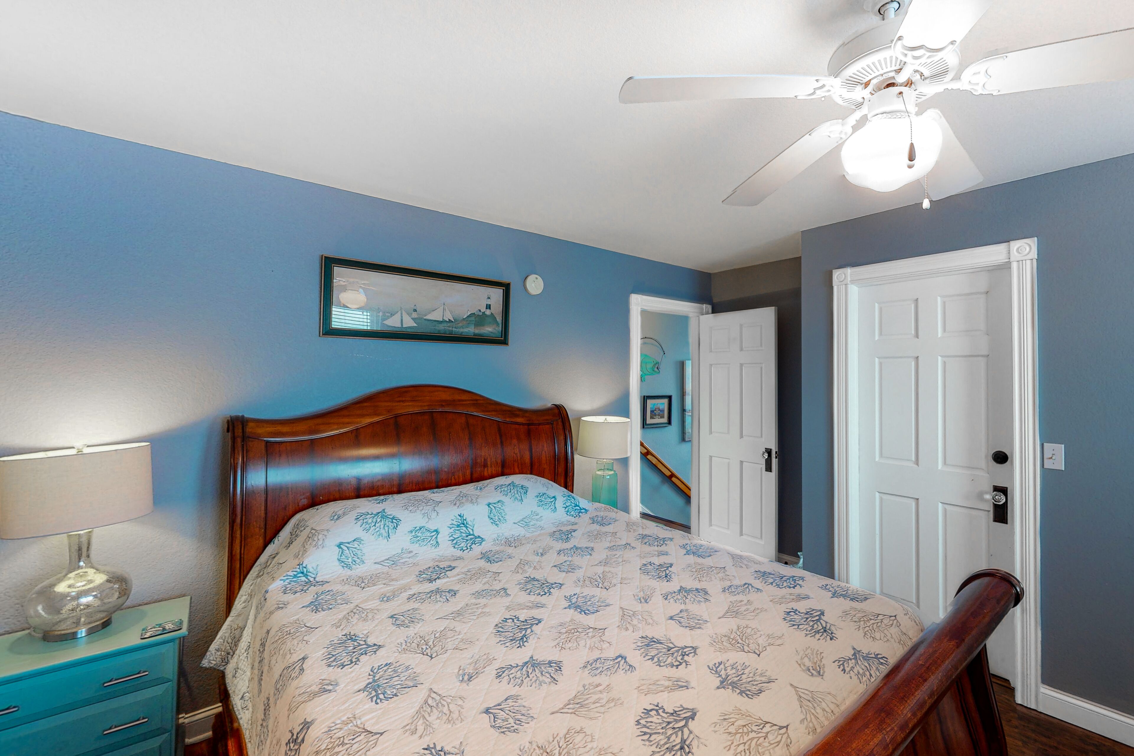 A bedroom with a wooden bed, a nightstand with a lamp, a ceiling fan, and a door leading to another room.