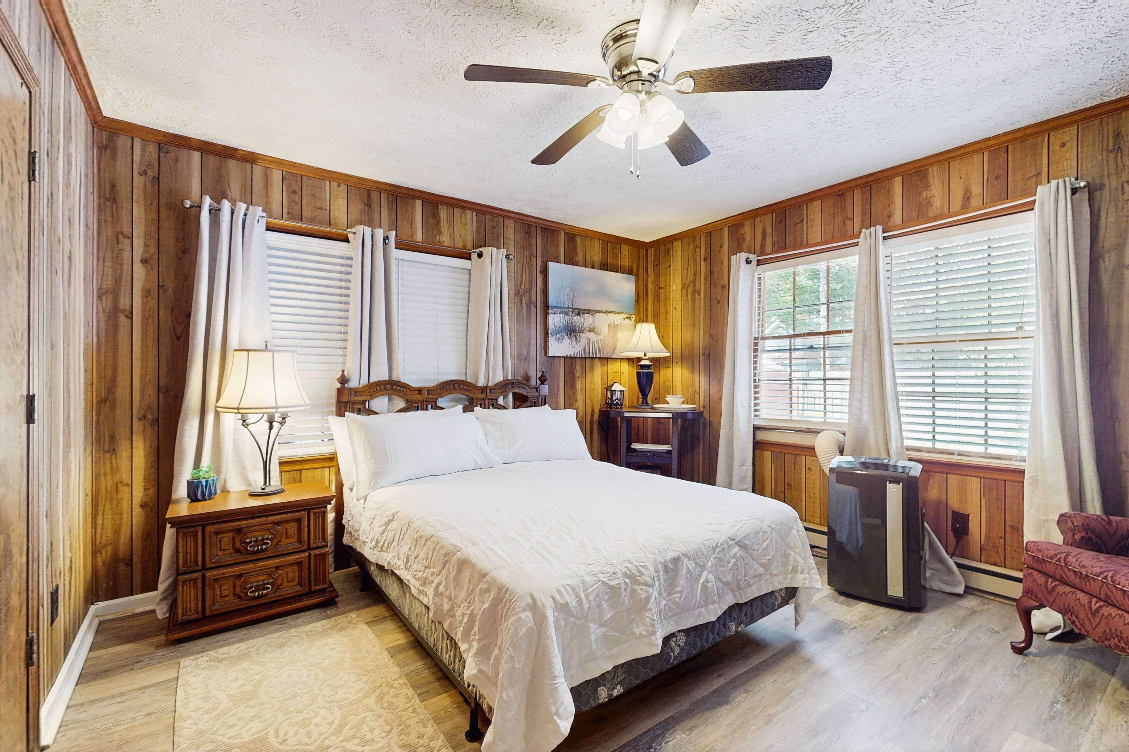 A bedroom with a wooden paneled wall, a bed with white bedding, a nightstand with a lamp, a dresser, a red armchair, and a ceiling fan.