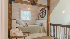 Un dormitorio tipo loft con techo inclinado, estructura de vigas de madera, un ventanal grande, una cama con almohadas, una silla y una rueda decorativa.