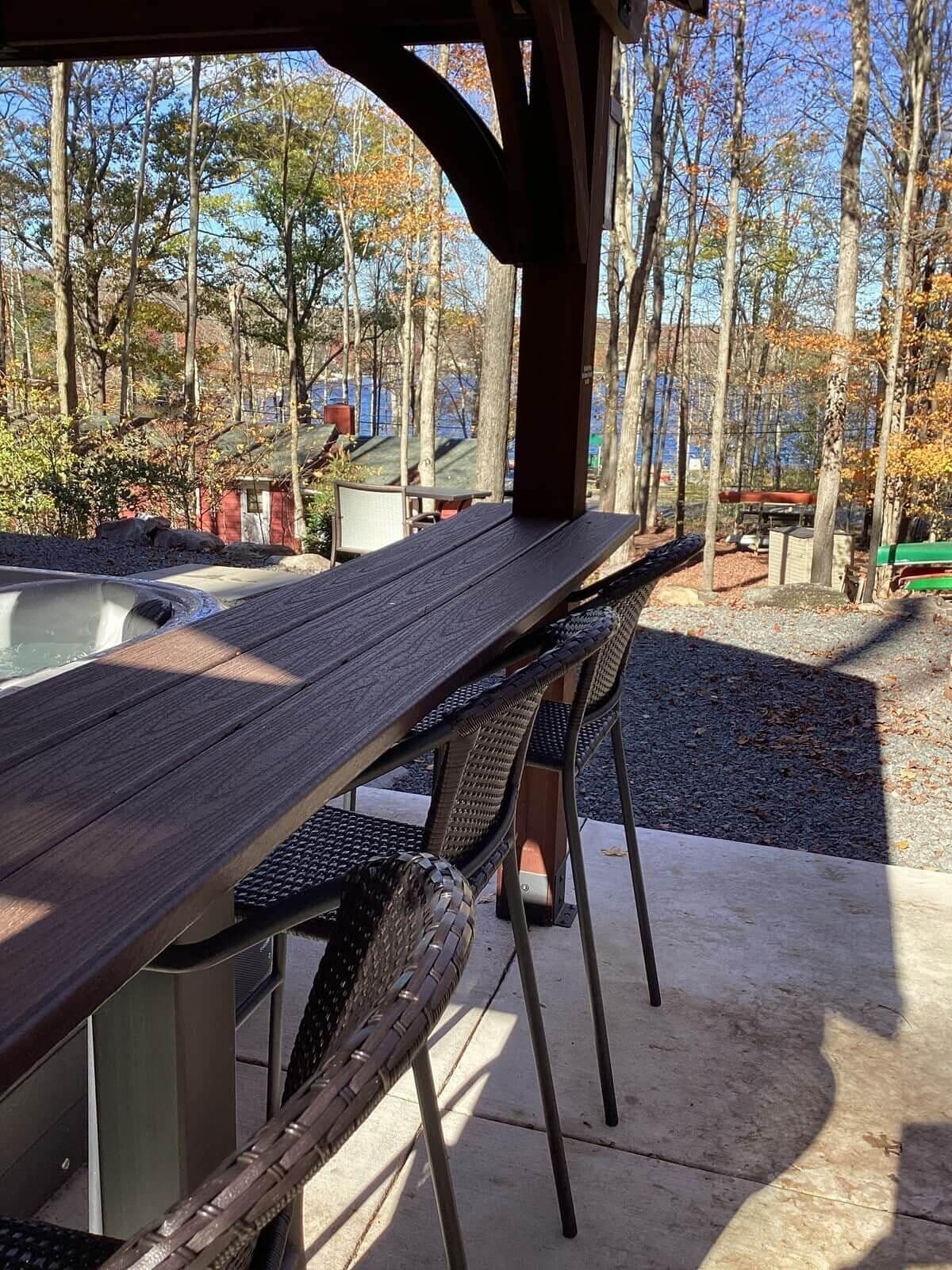 An outdoor patio with a long wooden table and chairs, surrounded by trees and a view of a body of water.