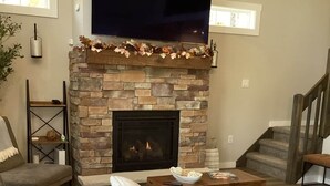 Una sala de estar con chimenea de piedra, un televisor de pantalla plana montado encima y una escalera a la derecha.