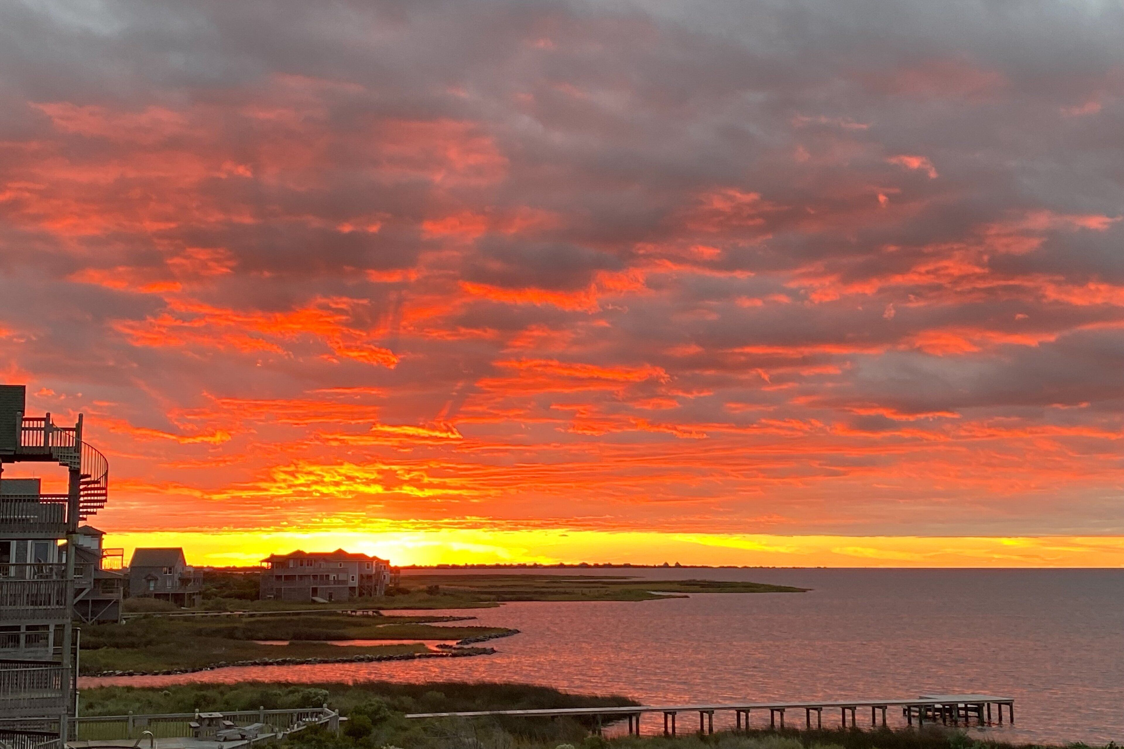 Une vue sur le coucher de soleil en bord de mer, avec une jetée en bois s’étendant dans l’eau, entourée de bâtiments et de zones marécageuses.