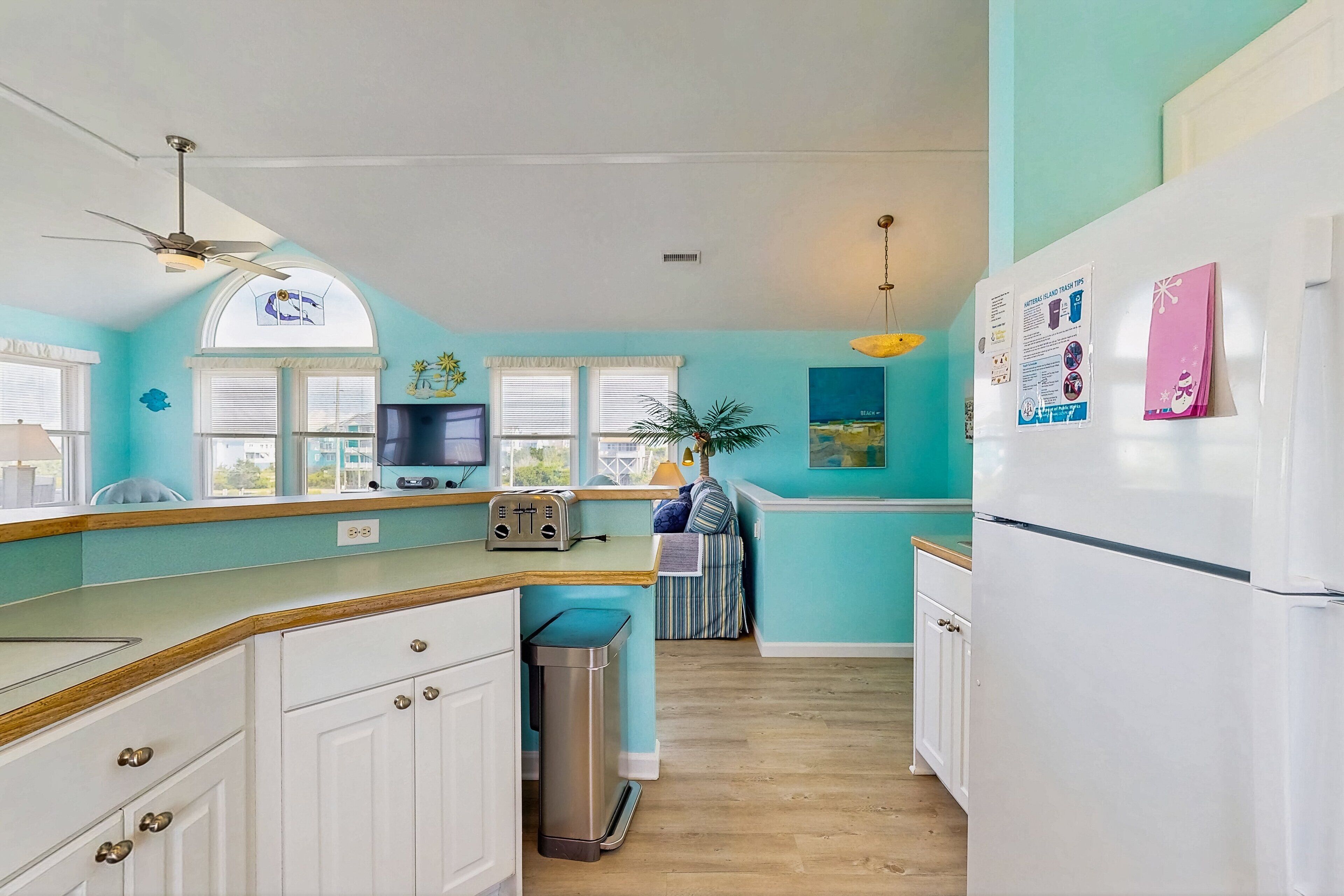 A kitchen with a toaster, a trash bin, a refrigerator, and a window with blinds.