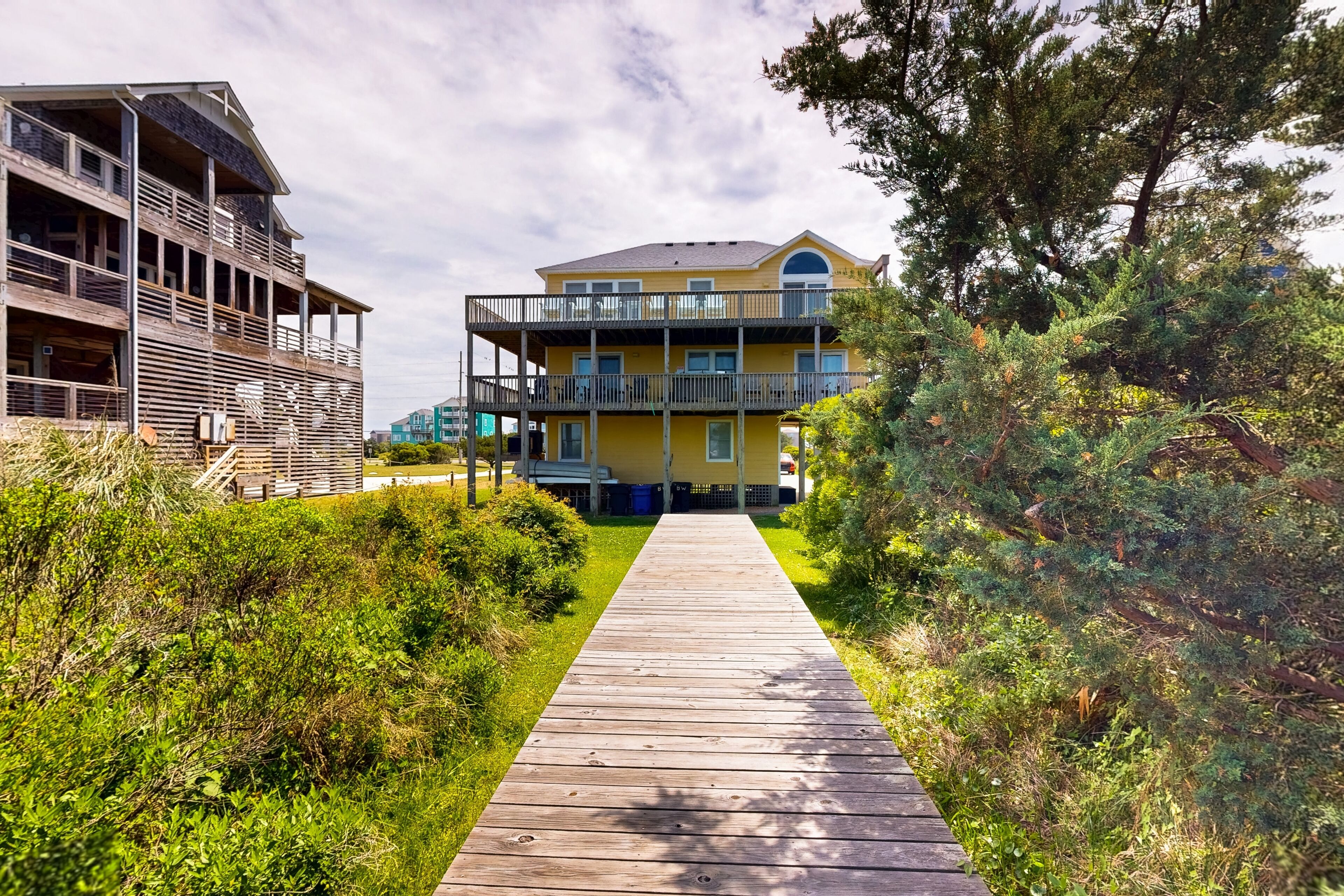 A wooden pathway leading to a yellow building with balconies, surrounded by greenery.
