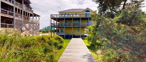 A wooden pathway leading to a yellow building with balconies, surrounded by greenery.
