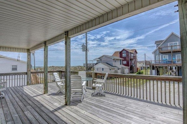 A wooden deck with lounge chairs, a covered area, and a view of houses and beach in the background.