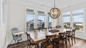 A spacious dining area with a large wooden table and multiple chairs, large windows offering a view of buildings, and a high ceiling with a central light fixture. - Sunny Daze: Soundview, Private Pool, Elevator (Ocean Isle Beach)