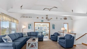 Een ruime woonkamer met een grote hoekbank, een salontafel en twee fauteuils. Er hangen decoratieve wanddecoraties en er is een plafondventilator.