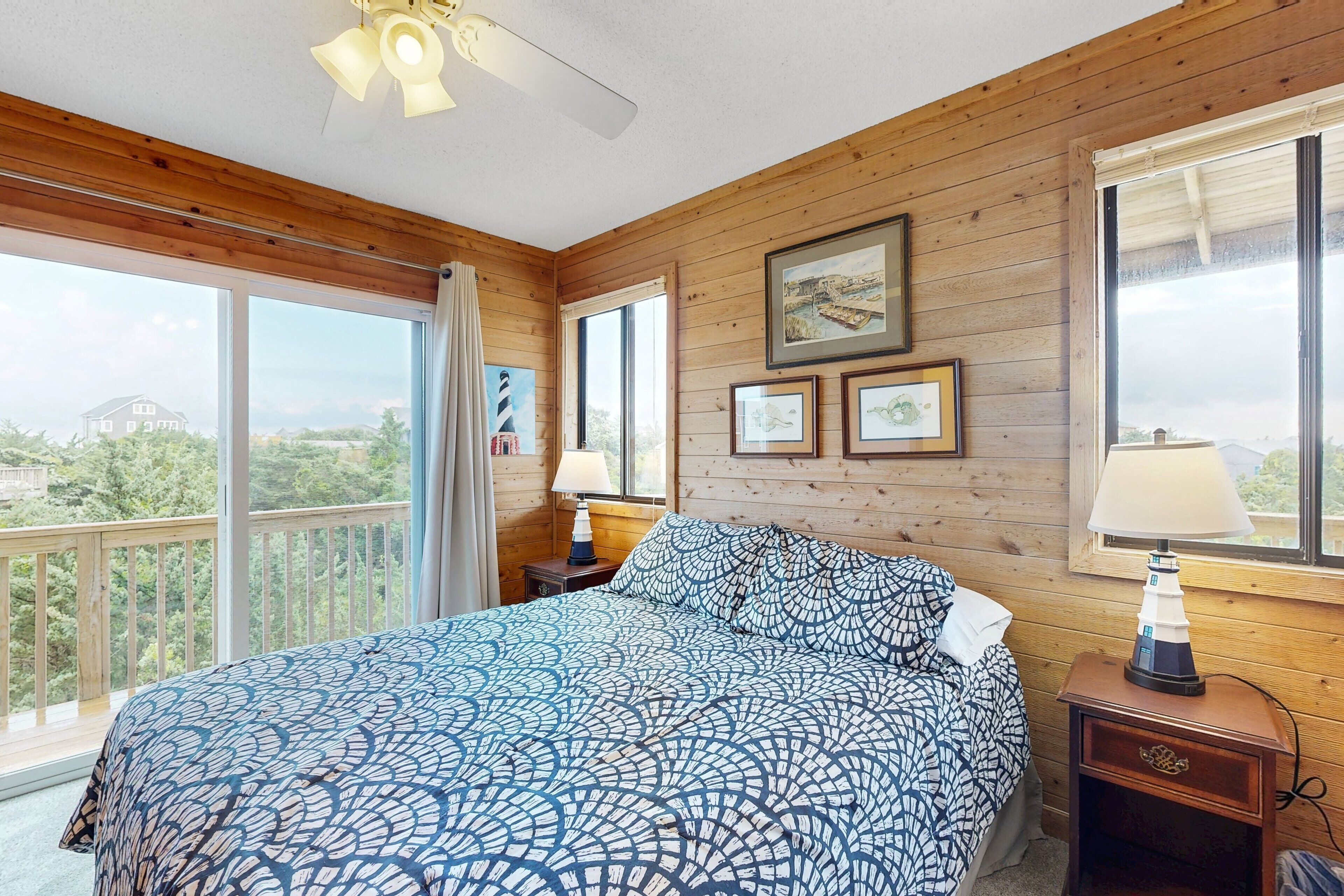 A bedroom with a large bed, wooden walls, and a balcony with a view of greenery and a distant building.
