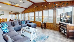 A spacious living room with a large sectional sofa, a coffee table, and a television. The room features wooden walls and large windows. - Attitude Adjustment: Dog-friendly, ocean/sound views, shared pool (Avon)
