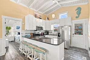 A modern kitchen with white cabinetry, a central island with stools, and a view through the door. - sandBar cottaGe | Private Pool & Ocean Views | Stroll 1 Minute to Beach (Ocean Isle Beach)