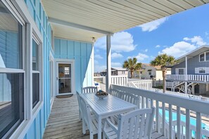 A covered deck with a white table and chairs, overlooking a pool and neighboring buildings. - Starfish Cottage | Private Pool & Ocean Views | Stroll 1 Minute to Beach (Ocean Isle Beach)