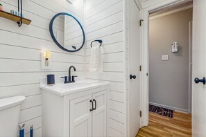 Une salle de bain avec un meuble-lavabo blanc, un miroir rond, une serviette accrochée au mur et une porte donnant sur une autre pièce.