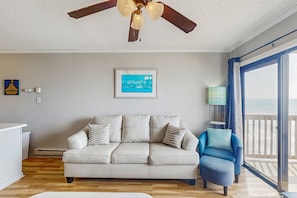 Un salon avec un canapé beige, un fauteuil bleu, un ventilateur de plafond, un tableau encadré et une vue sur l’océan par de grandes fenêtres.
