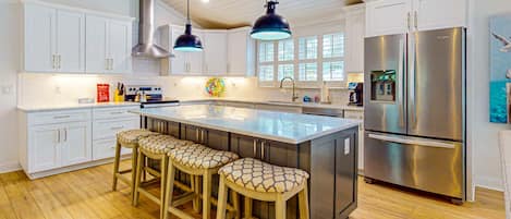 A modern kitchen with white cabinets, a stainless steel refrigerator, wooden bar stools, and pendant lighting.