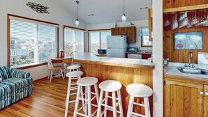 A modern kitchen with wooden cabinets, a white countertop, and a dining area with a striped sofa and white stools. - Oceanfront house w/private hot tub, balcony, washer/dryer, WiFi, central AC (Waves)