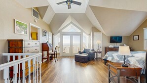 A spacious loft bedroom with a high ceiling, a ceiling fan, a large bed, a desk, a chair, and a view through a glass door to the outside.